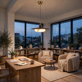 A modern mid-century style pendant light with a warm glowing bulb hangs in a cozy, contemporary home office/lounge area with large windows, wooden furniture, and plants.