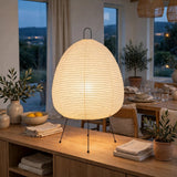 A glowing paper lantern table lamp with black metal tripod legs sits on a wooden console table in a warmly lit dining room.
