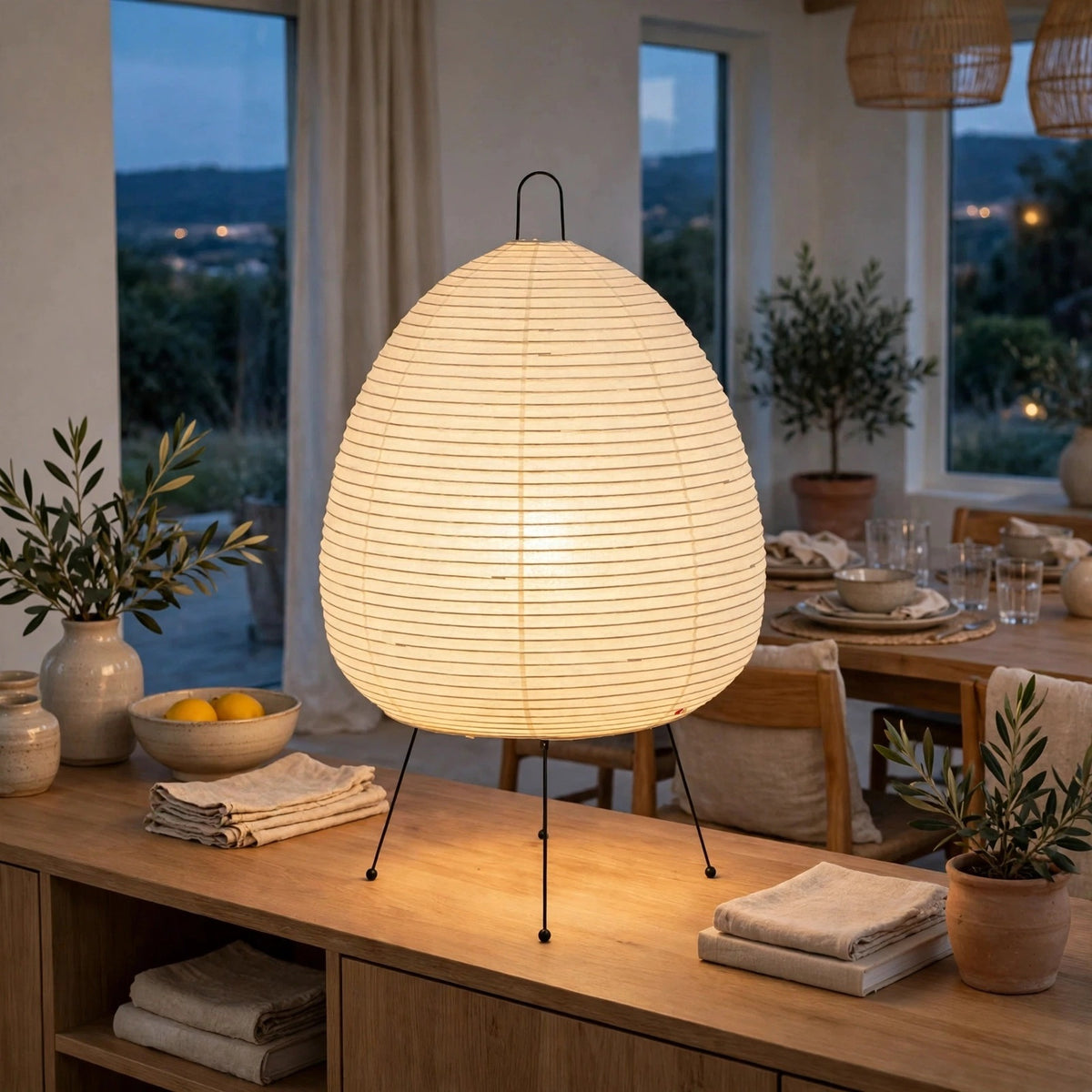 A glowing paper lantern table lamp with black metal tripod legs sits on a wooden console table in a warmly lit dining room.