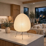 A large, egg-shaped table lamp with a ribbed white paper or fabric shade and three thin black metal legs, glowing warmly on a kitchen island in a modern, upscale living room.