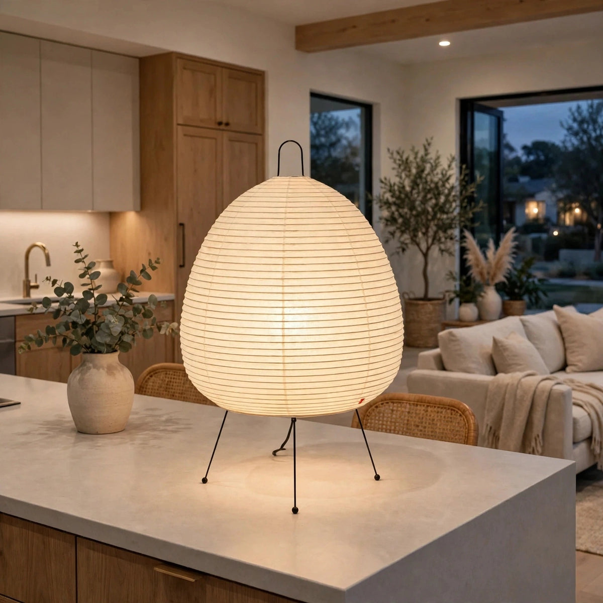 A large, egg-shaped table lamp with a ribbed white paper or fabric shade and three thin black metal legs, glowing warmly on a kitchen island in a modern, upscale living room.