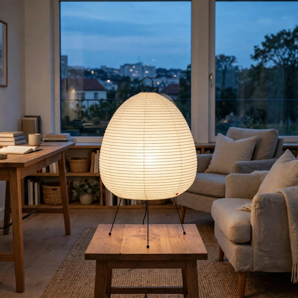 A glowing teardrop-shaped paper lantern table lamp with black metal legs sits on a wooden table in a cozy, modern living room at dusk.