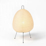 A modern table lamp with a ribbed, egg-shaped beige paper shade on a black metal tripod base, photographed against a clean white background.