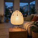 A glowing table lamp with a ribbed egg-shaped shade and thin black legs sits on a wooden side table in a cozy living room at dusk.