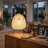 A warm, glowing table lamp with a ribbed, egg-shaped beige shade and thin black metal legs, set on a wooden sideboard in a beautifully decorated dining room at dusk.