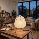 A modern egg-shaped table lamp with a warm white ribbed paper shade and black metal legs, placed on a wooden coffee table in a cozy, modern living room at dusk.