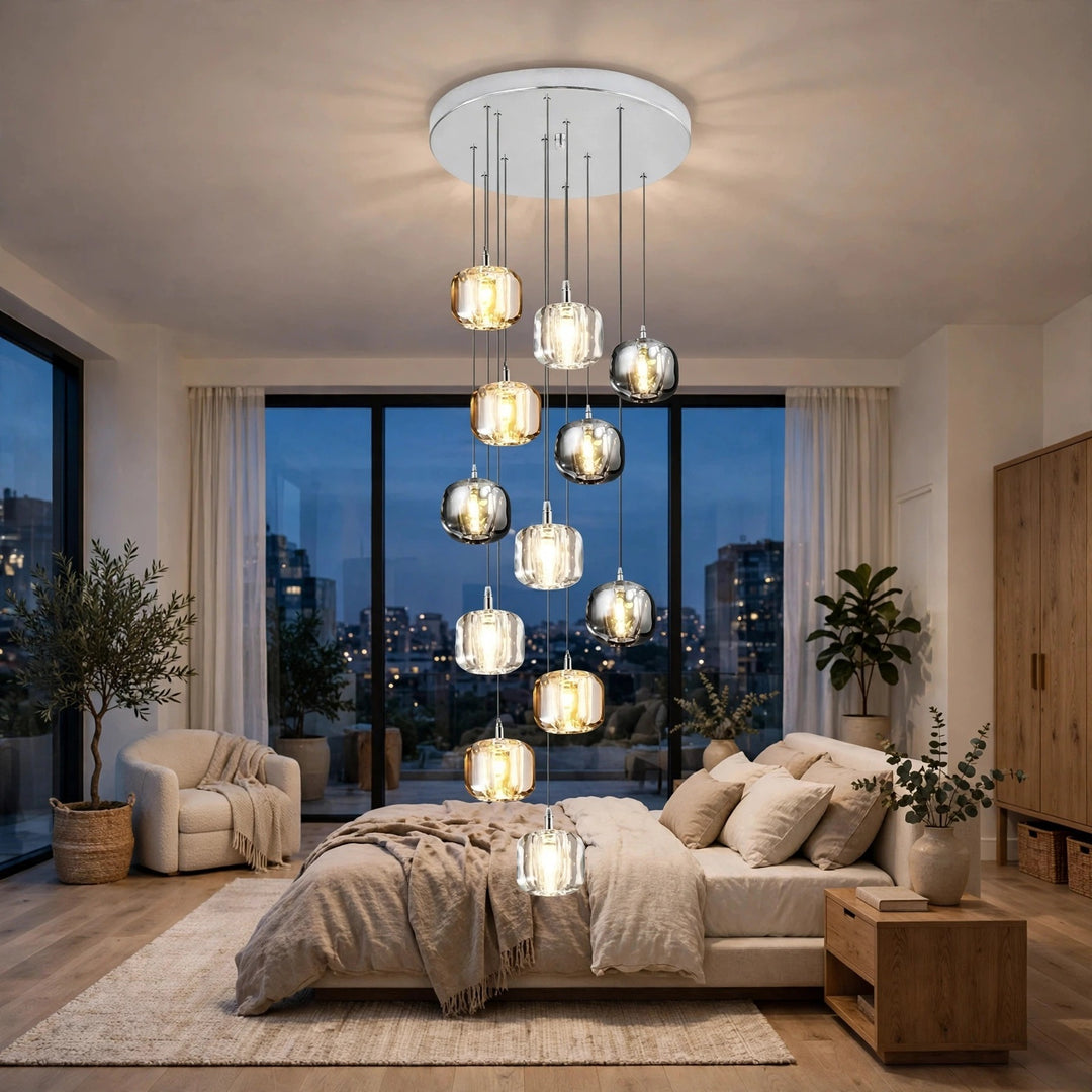 A modern multi-light chandelier with clear glass cylindrical and spherical pendant shades hangs over a bed in a contemporary bedroom with large windows, city views at dusk, beige bedding, and wood furniture.
