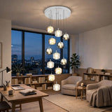 A modern multi-light pendant chandelier with clear glass cylindrical globes and warm white bulbs hanging from a white ceiling canopy in a stylishly furnished living room with a desk, armchairs, and large windows overlooking a city skyline at dusk.