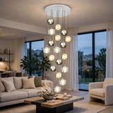 A modern multi-light chandelier with clear glass cylindrical shades and warm glowing bulbs, suspended from a white ceiling in a contemporary living room with a sofa, coffee table, and large windows.