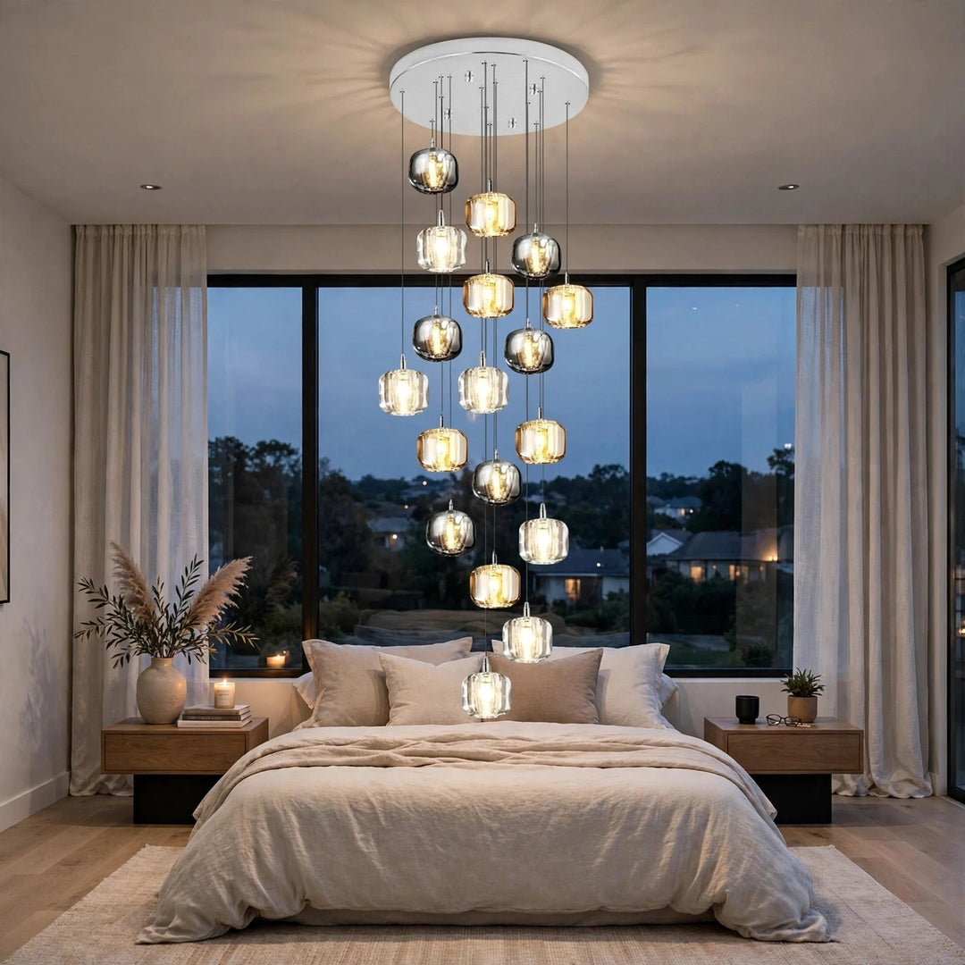 A modern multi-light chandelier with clear glass cylindrical shades and warm white bulbs, suspended from a circular white ceiling mount above a bed in a styled bedroom.