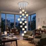 A modern multi-light chandelier with clear glass and metallic spherical shades hangs in a stylishly furnished living room with large windows overlooking a city skyline at dusk.