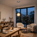 A single-light amber crystal chandelier glows above a wooden desk beside cream armchairs and large windows at dusk.