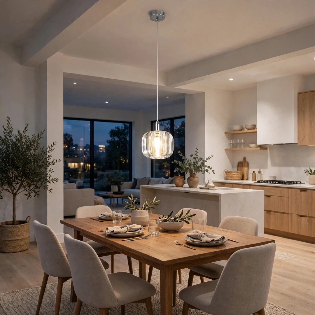A single-light clear crystal chandelier glows above a wooden dining table beside an open kitchen with light wood cabinetry.