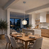 A single-light clear crystal chandelier glows above a wooden dining table beside an open kitchen with light wood cabinetry.