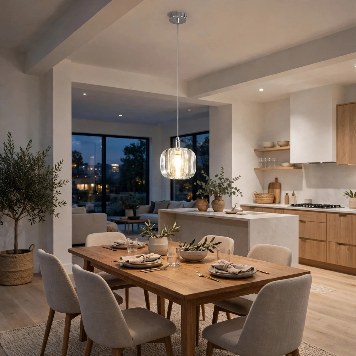 A single-light clear crystal chandelier glows above a wooden dining table beside an open kitchen with light wood cabinetry.