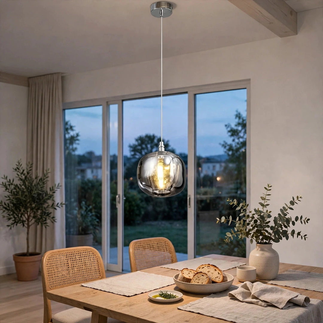 A single-light smoky gray crystal chandelier hangs above a wooden dining table with woven chairs beside tall garden windows.