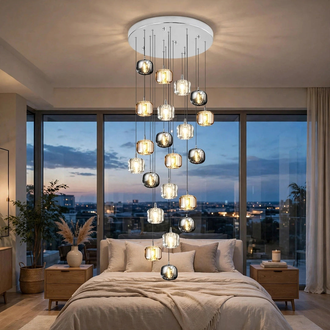 A modern multi-tier chandelier with clear glass spherical pendant lights and warm glowing bulbs, hanging from a white ceiling in a contemporary bedroom with large windows overlooking a city skyline at dusk.
