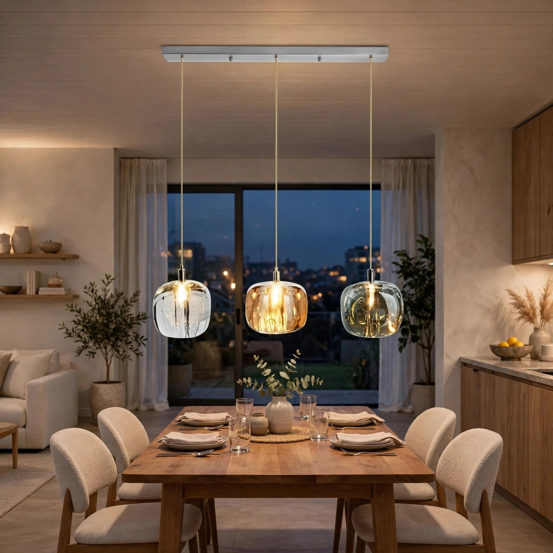 A modern 3-light linear ceiling pendant chandelier with clear glass globes and warm glowing bulbs, hanging over a wooden dining table in a contemporary dining room.