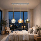 Linear chandelier with three clear, amber, and smoky crystal shades hangs above a white bed beside large windows overlooking the city at dusk.