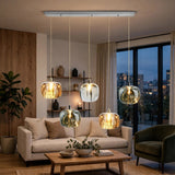 A modern 5-light linear ceiling pendant chandelier with clear glass bubble shades glowing with warm yellow light, installed in a contemporary living room with a sofa, coffee table, and large window overlooking the evening city skyline.