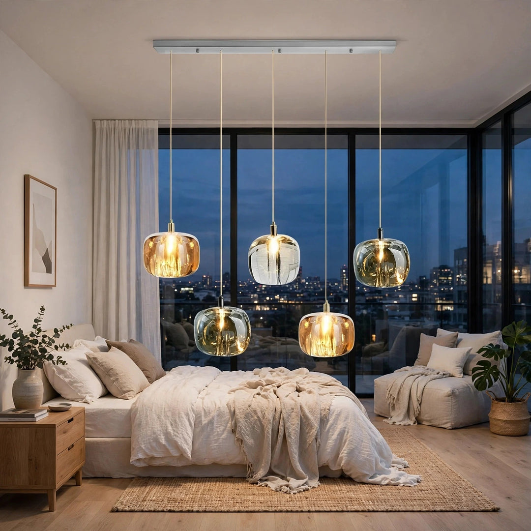 A five-light linear chandelier with amber, clear, and smoky crystal shades hangs above a cream bed beside corner windows.