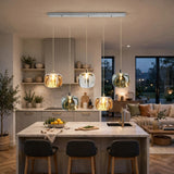 A five-light linear chandelier with amber, clear, and smoky crystal shades illuminates a pale kitchen island with three dark stools.