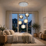 A modern multi-light pendant chandelier with five smoky glass shades hangs over a neatly made bed in a softly lit bedroom.