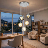 A modern multi-light pendant chandelier with five clear glass globes and warm white bulbs hanging in a stylishly lit living room with a desk, chairs, and bookshelves.