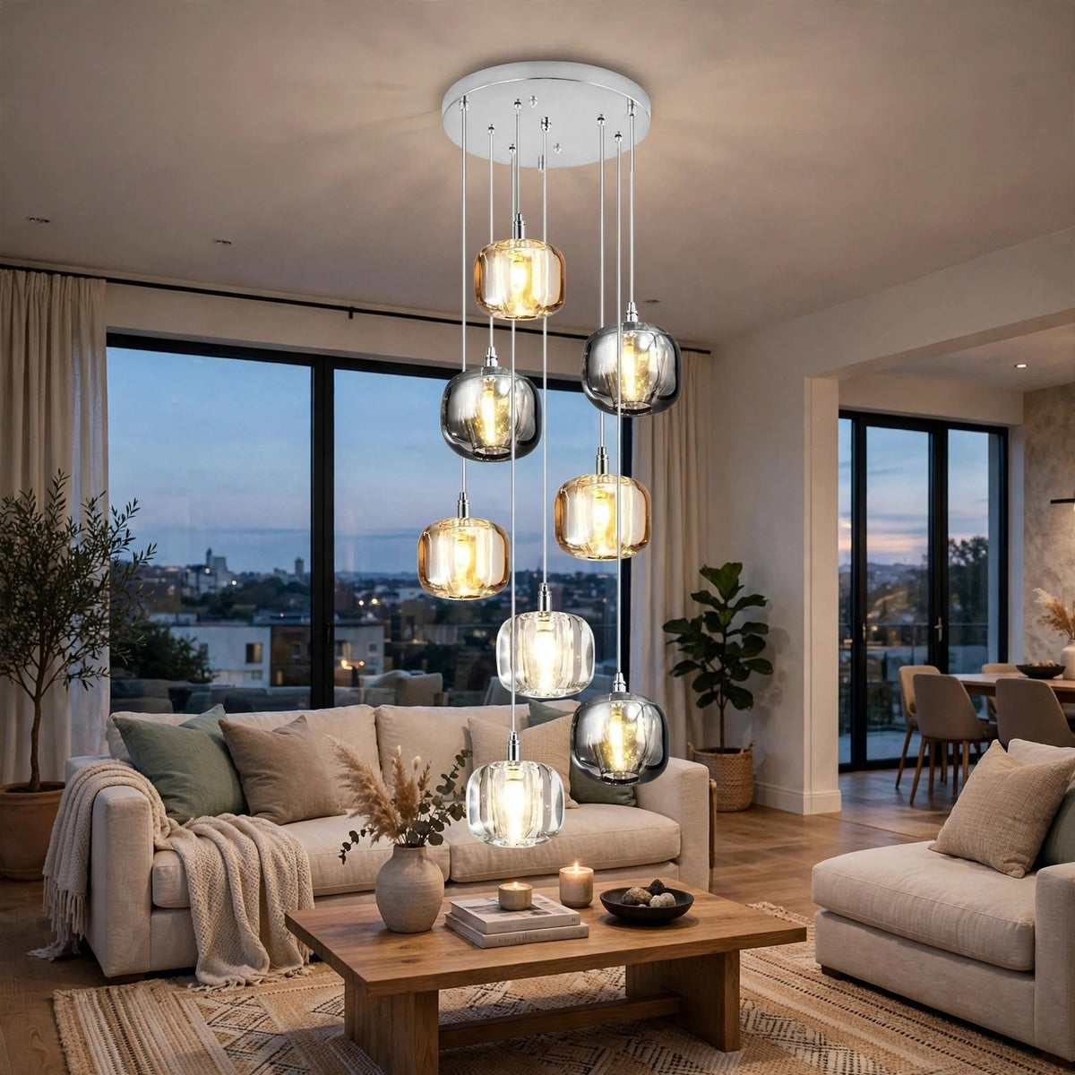 A modern multi-light pendant chandelier with clear glass spherical shades and warm glowing bulbs, hanging in a contemporary living room with beige sofas, a wooden coffee table, and large windows overlooking a city skyline at dusk.