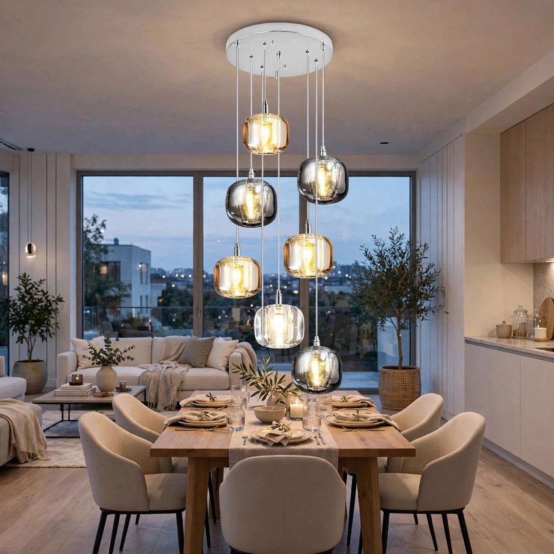 A modern multi-light pendant chandelier with six glass shades in silver, smoked gray, and clear, glowing with warm light in a contemporary dining room.