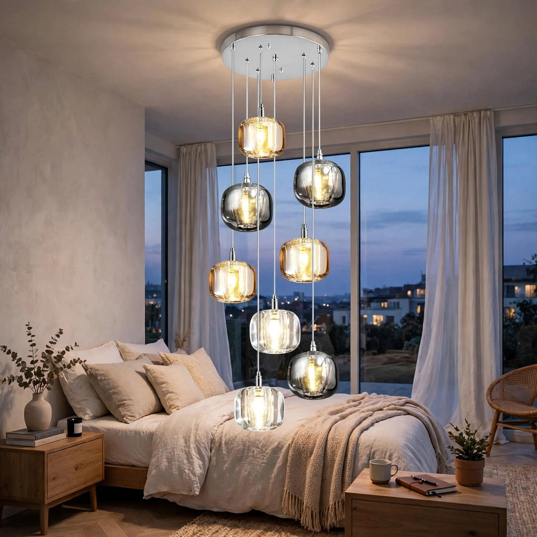 A modern multi-light chandelier with seven hanging smoked glass globes and warm glowing bulbs, installed in a bedroom above a bed with beige bedding and large windows showing a dusk view.