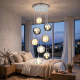 A modern multi-light chandelier with seven hanging smoked glass globes and warm glowing bulbs, installed in a bedroom above a bed with beige bedding and large windows showing a dusk view.