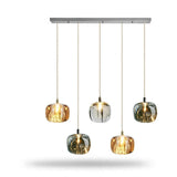 A linear chandelier has five illuminated amber, clear, and smoky grey shades at alternating heights beneath a rectangular silver canopy against white.