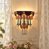 A decorative wall sconce with a dark brown top band, peach and orange teardrop-shaped glass drops, and blue-green hanging stems, mounted on a cream paneled wall between sheer curtains.