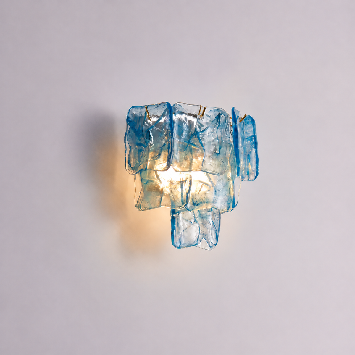A modern wall sconce light fixture made of stacked translucent blue glass blocks, casting a warm yellow glow against a plain light gray wall.