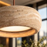 A close-up of a modern pendant light with a textured beige travertine stone shade, suspended by a thin copper-colored cord against a softly blurred interior background.