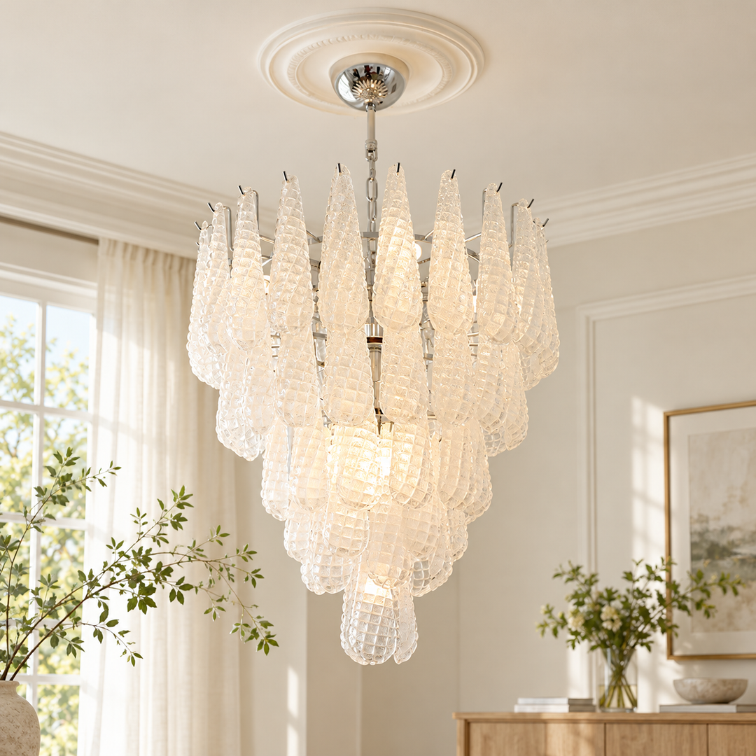 A modern multi-tiered chandelier with textured, translucent teardrop-shaped glass shades and a polished silver-toned metal frame, hanging from a decorative ceiling medallion in a bright, elegant room.