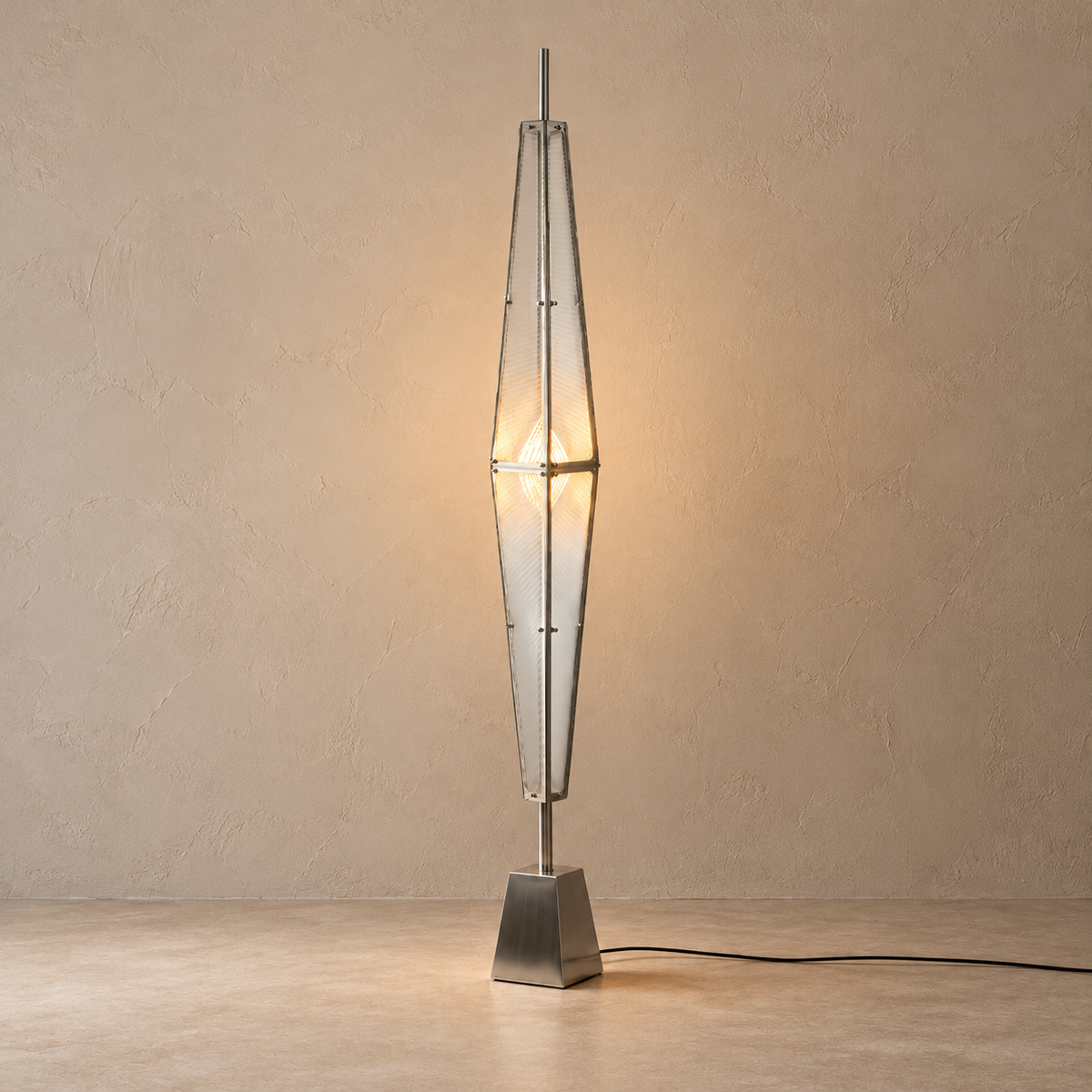 A modern geometric floor lamp with a silver metal frame and clear glass panels, illuminated with a warm light, standing on a square metallic base against a textured beige wall.