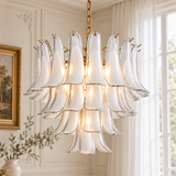 A modern multi-tiered chandelier with translucent white glass petal-like shades and gold-toned metal accents, hanging in a bright, elegant room with white walls and a window.