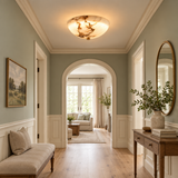 A beautifully styled hallway with sage green walls, white wainscoting, a warm wood floor, and a glowing oval ceiling light. On the left is a beige upholstered bench, and on the right is a wooden console table with a vase of greenery and an oval mirror.