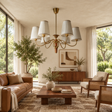 A beautifully styled mid-century modern living room featuring a brass chandelier with white pleated drum shades, a brown leather sofa, a wooden coffee table, and large windows looking out onto green trees.