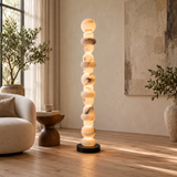 A tall, sculptural floor lamp made of stacked glowing stone-like spheres on a black base, placed in a softly lit living room beside a beige armchair, small wooden side table, and potted plants.