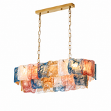 Linear chandelier with layered multicolour art glass panels in blue, amber, and coral hanging from gold chains on a white background.