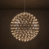A spherical geometric wire chandelier pendant light with many small illuminated bulbs, hanging from a thin cord against a dark brown wall and ceiling.