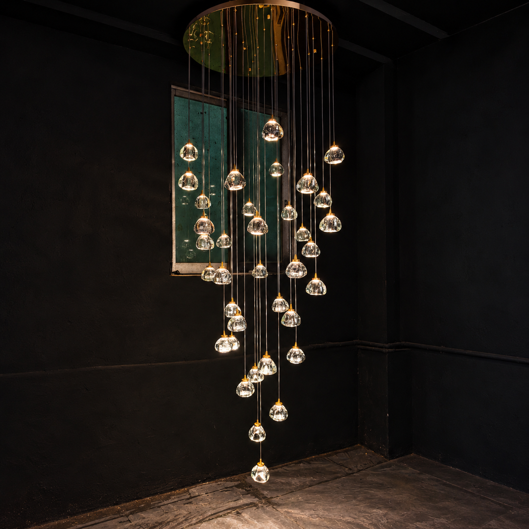 A modern multi-light chandelier with clear glass teardrop pendant bulbs suspended from a circular gold-toned metal frame, hanging in a cascading waterfall pattern against a dark black wall.