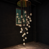 A modern multi-light chandelier with clear glass teardrop pendant bulbs suspended from a circular gold-toned metal frame, hanging in a cascading waterfall pattern against a dark black wall.