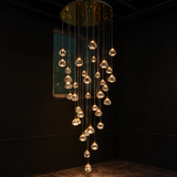 A modern multi-light chandelier with numerous clear glass teardrop pendant bulbs hanging from thin cables in a cascading arrangement, illuminated in a dark room corner.