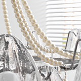 A close-up of a modern chandelier featuring a prominent strand of cream pearl beads draped over clear glass and textured silver metal arms.