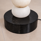 A close-up of a table lamp base featuring a round black stone pedestal and a stacked white stone sphere column, set on a beige tiled floor.