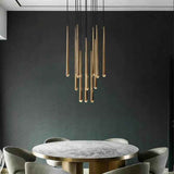 A modern multi-light linear chandelier with brushed gold rods and black cords, hanging above a round marble dining table with gray chairs against a dark gray wall.